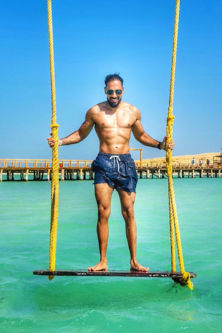 Bodybuilder am Strand Ein muskulöser Mann schwingt auf einer Schaukel über türkisfarbenem Wasser.